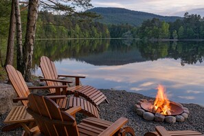 A lakeside setting with wooden Adirondack chairs and a campfire. - The Queen's Hut - Idyllic Log Cabin, originally built for Queen Mary (Aboyne)