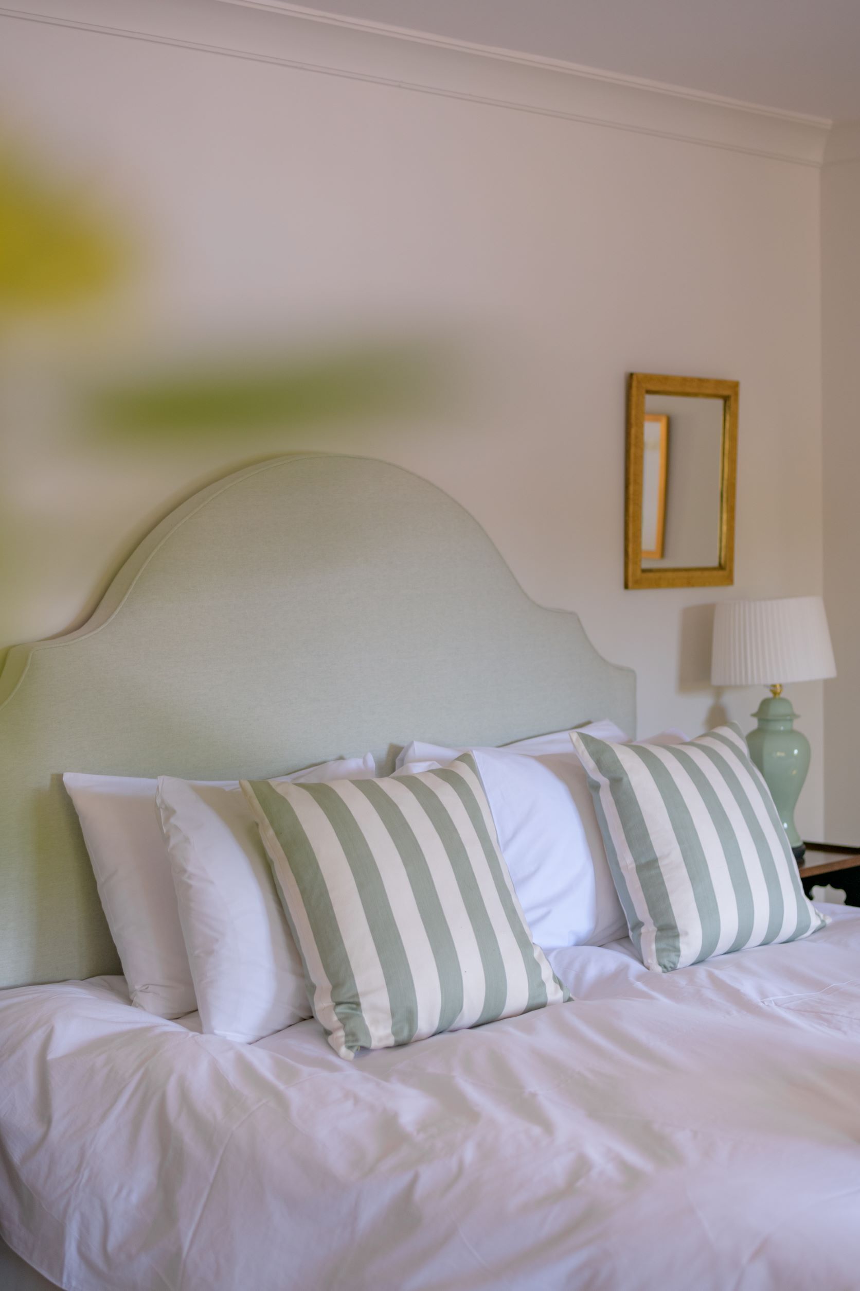 Un lit avec une literie blanche et des oreillers à rayures, une tête de lit verte, une table de chevet avec une lampe et un miroir encadré accroché au mur.