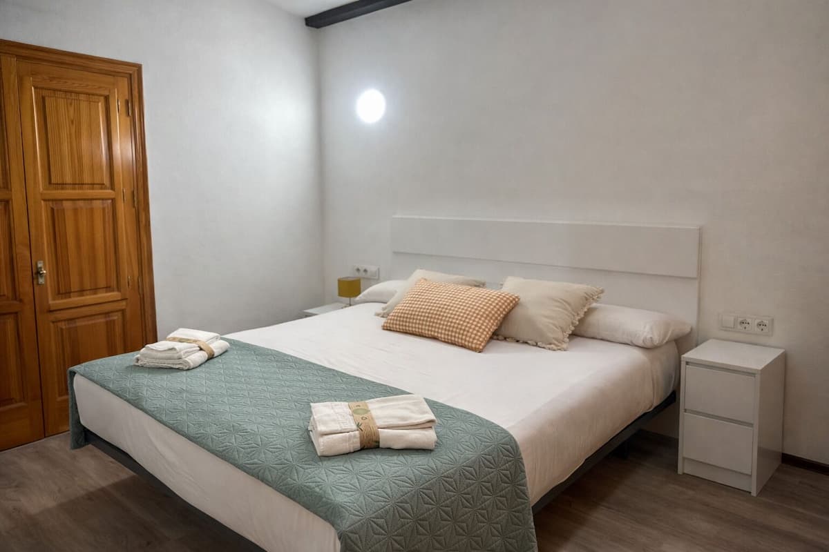 Un lit bien fait, recouvert d’une couette verte, avec deux oreillers et des serviettes pliées posées sur une table de chevet blanche.