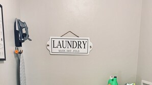 Uma lavanderia com uma placa indicando "LAUNDRY WASH. DRY. FOLD", uma máquina de lavar, uma secadora e um aspirador de pó.