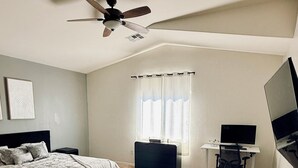 Quarto com cama, escrivaninha, cadeira, televisão, ventoinha no teto e janela com cortinas.