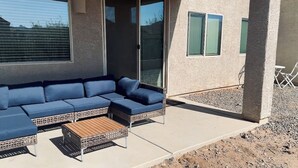 Uma área de estar coberta ao ar livre, com um sofá modular azul e uma mesa de centro de madeira.