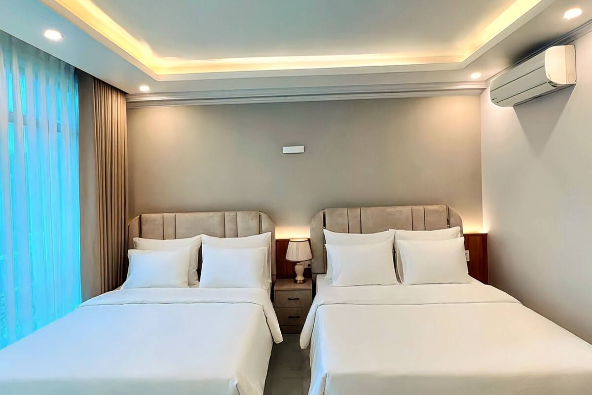 A hotel room with two beds, a nightstand, a lamp, and an air conditioning unit on the wall.