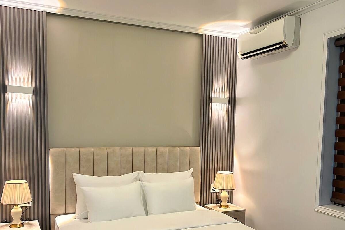 A modern hotel room with a large bed, two bedside tables with lamps, a ceiling light, and an air conditioning unit.
