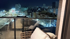 Un balcón con barandilla de vidrio, un sofá de mimbre con cojines y vistas a la ciudad de noche.