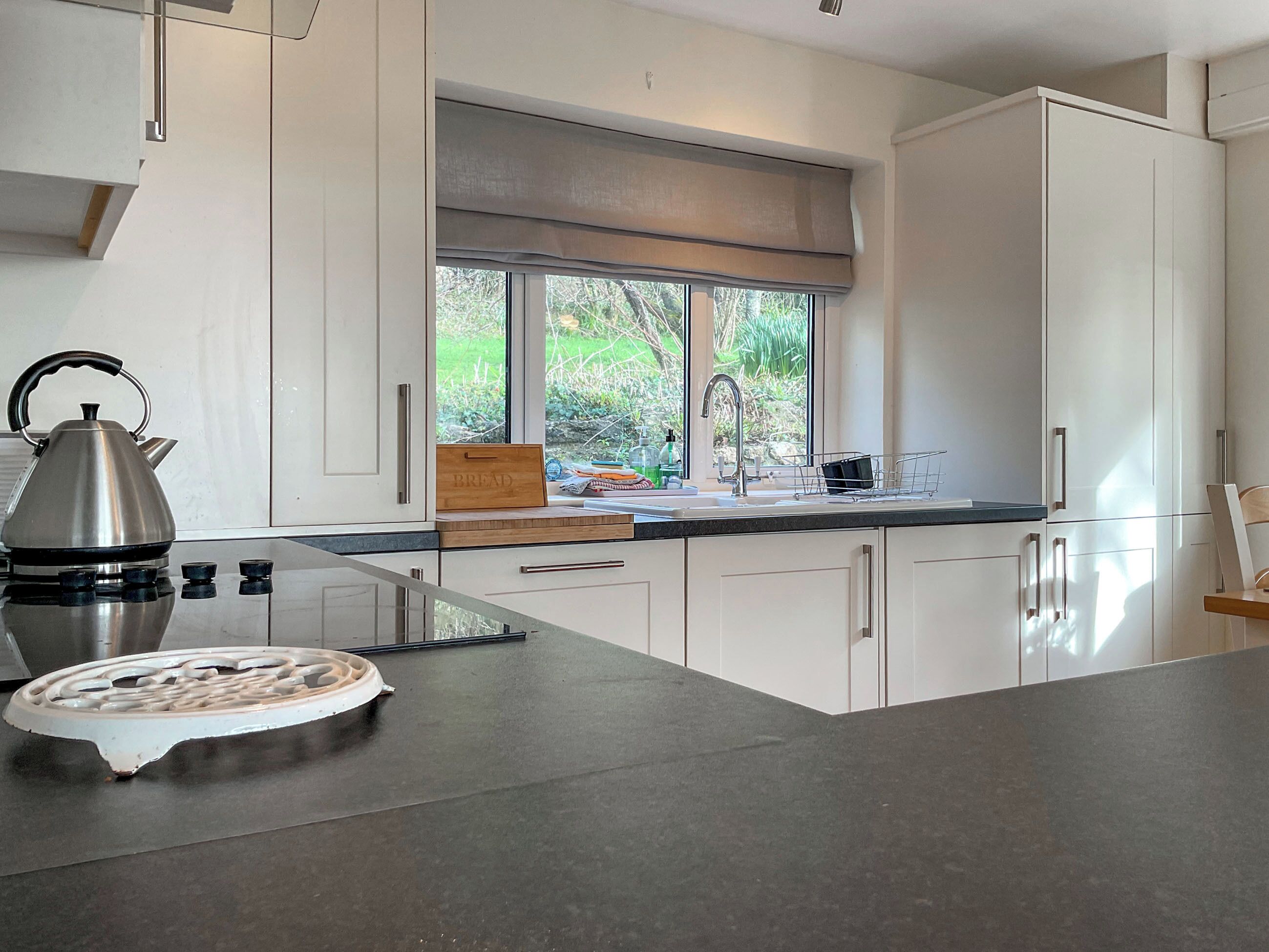 A modern kitchen with white cabinets, a stainless steel kettle, a bread box, and a window with a view of greenery.