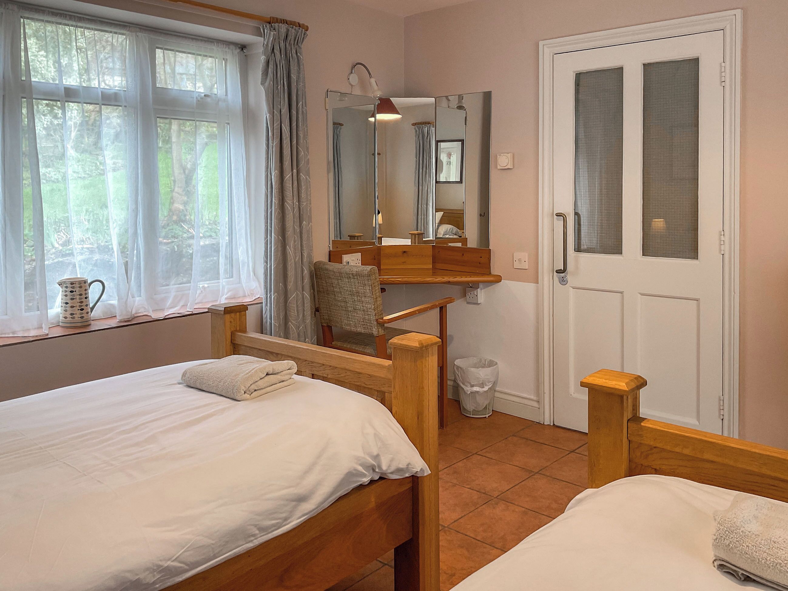 A bedroom with two wooden beds, a window with sheer curtains, a mirror, a wooden chair, and a door with a frosted glass panel.