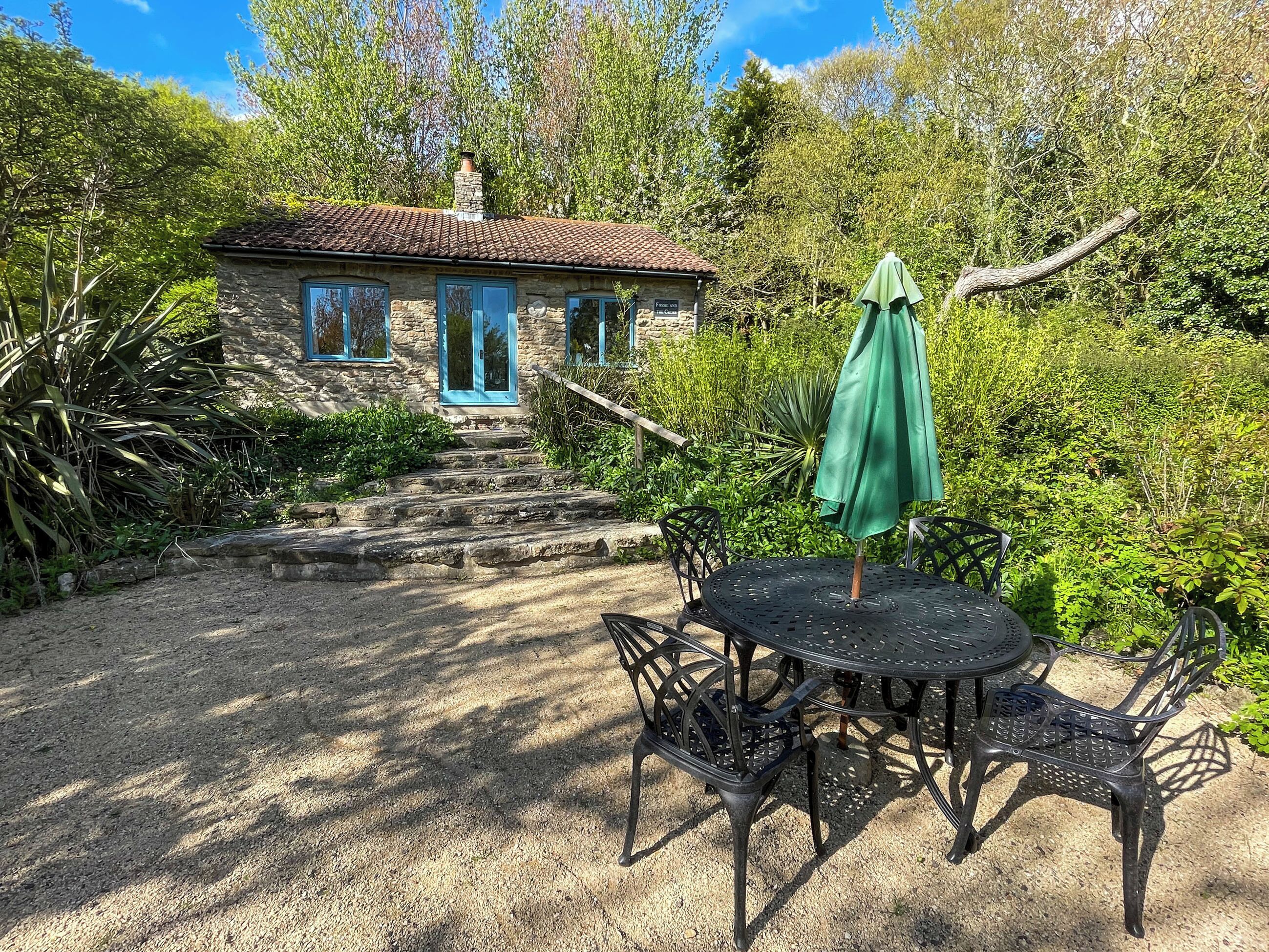 A stone building with a tiled roof, a green umbrella, and a table with chairs in a garden setting.