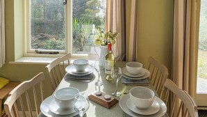 Una mesa de comedor servida con platos, tazones y copas de vino blancos, con una botella de vino y un florero con flores.
