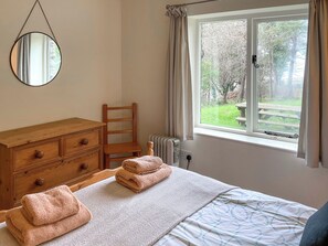 A bedroom with a wooden dresser, a chair, a bed with a patterned bedspread, a window with a view of greenery, and a round mirror on the wall.