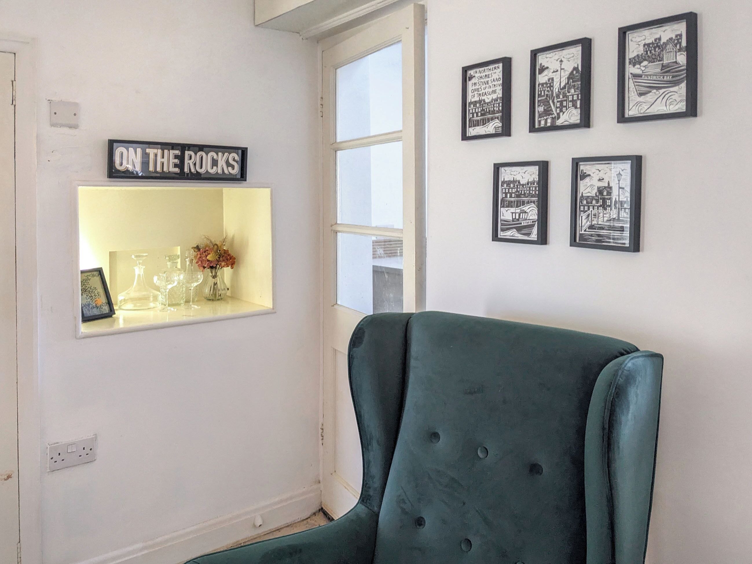 A room with a green armchair, a shelf with glassware and a sign reading 'ON THE ROCKS', and framed black and white pictures on the wall.