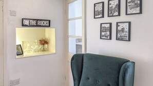 Una stanza con una poltrona verde, uno scaffale con bicchieri e un cartello con su scritto 'ON THE ROCKS', e fotografie in bianco e nero appese al muro.