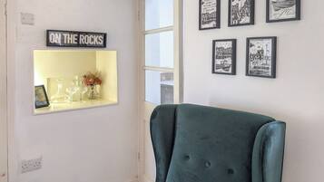 A room with a green armchair, a shelf with glassware and a sign reading 'ON THE ROCKS', and framed black and white pictures on the wall.