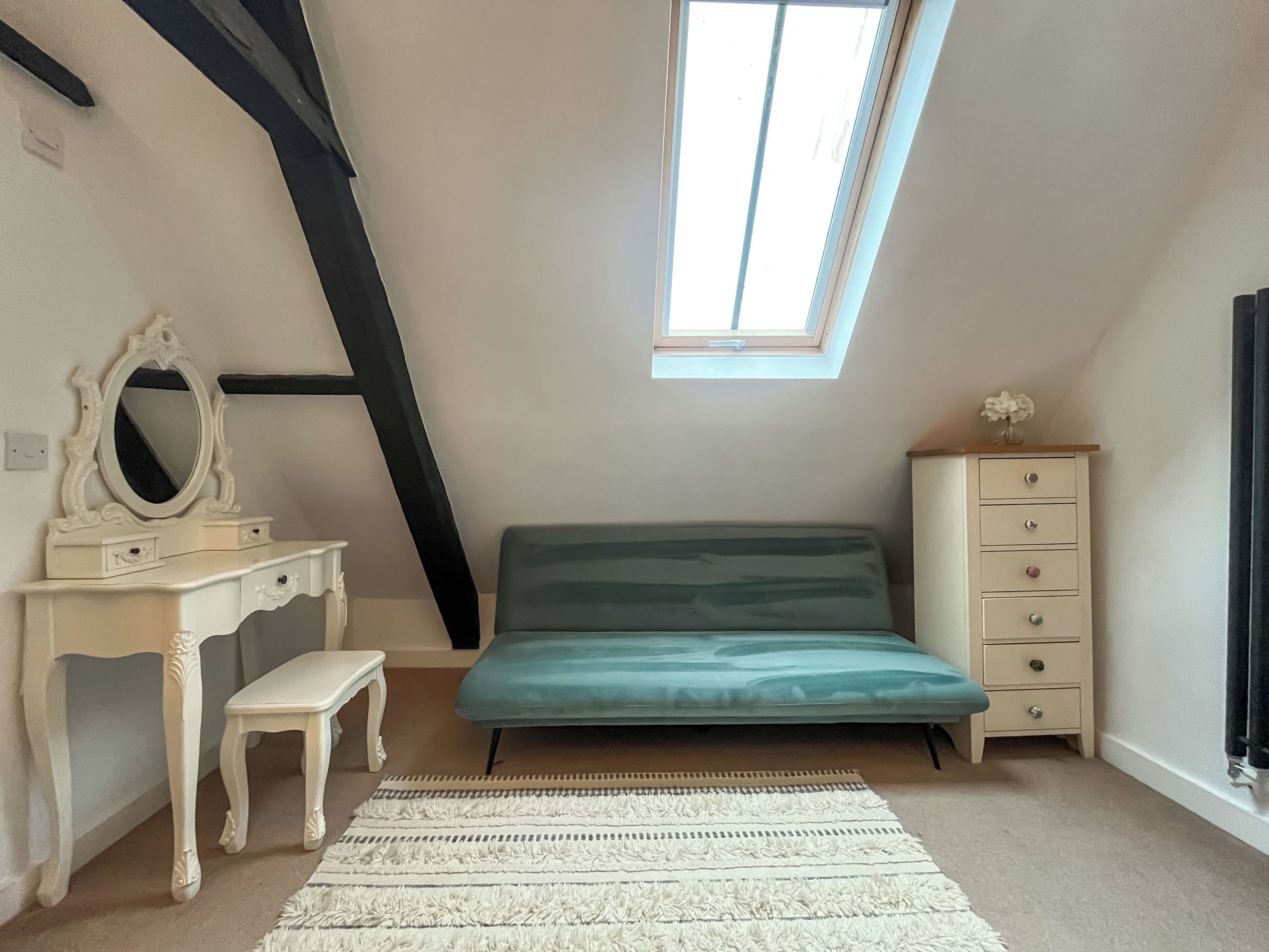 Ein Schlafzimmer mit schrägem Dach, einem Oberlicht, einem Bett, einem Schminktisch, einer Kommode und einer kleinen Bank.