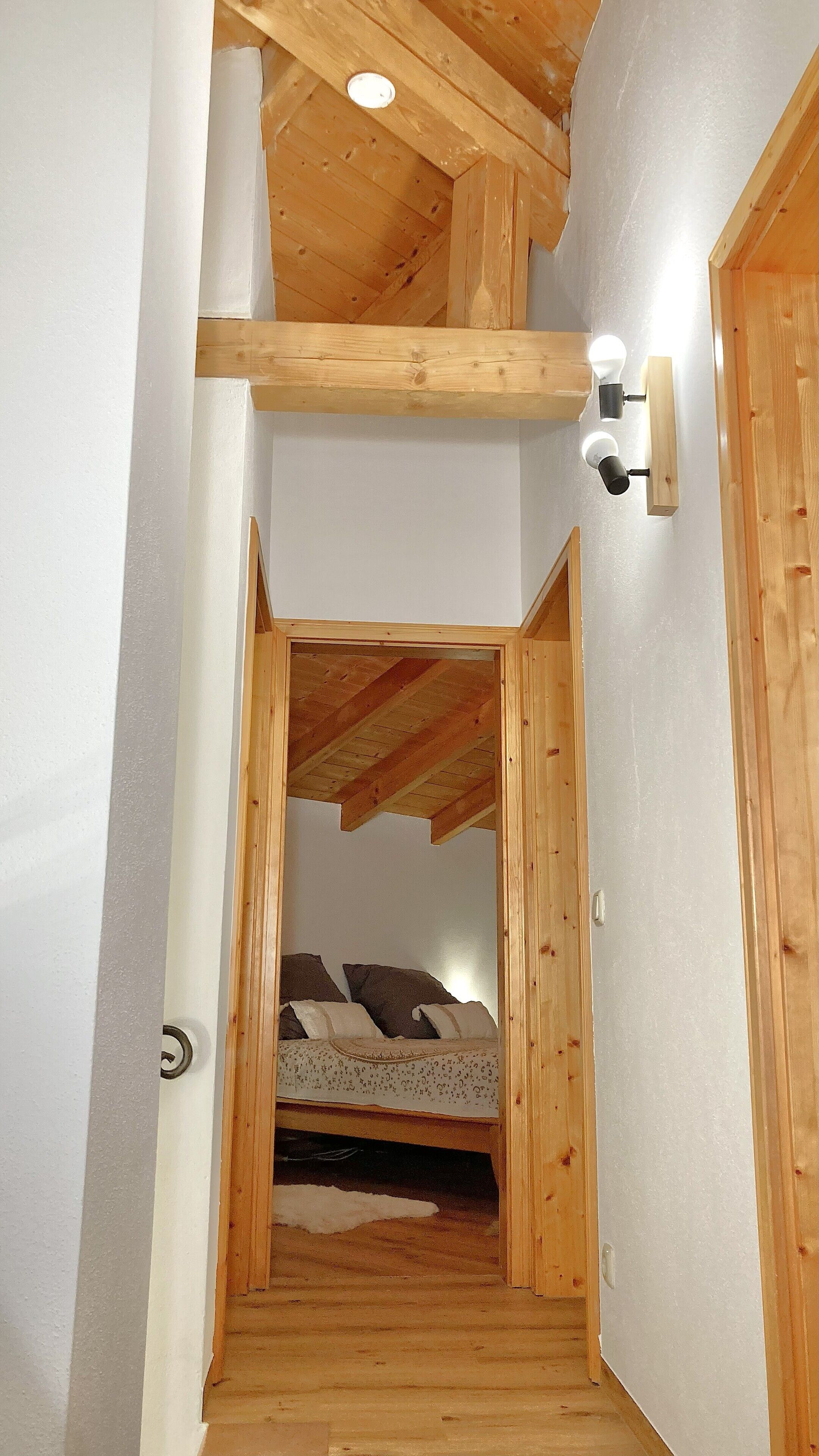 Ein schmaler Holzboden führt zu einem Schlafzimmer mit schrägen Wänden und einem Bett mit Kissen.