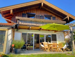 Ein zweistöckiges Holzhaus mit einem Balkon, einer überdachten Terrasse mit Tisch und Stühlen und einem grünen Sonnenschirm.
