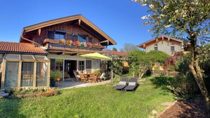 Une maison en bois dotée d’un balcon, d’une terrasse couverte et d’un jardin aménagé avec du mobilier d’extérieur.