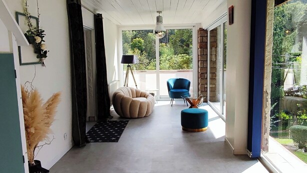 Een modern interieur met een hoge plafond, een centraal lichtarmatuur, een bank, een stoel en uitzicht door grote ramen.