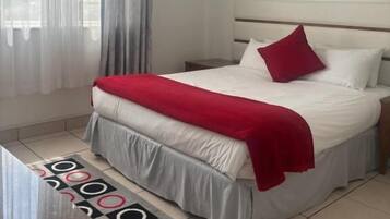 A hotel room with a bed, a ceiling fan, an air conditioner, and a window with sheer curtains.