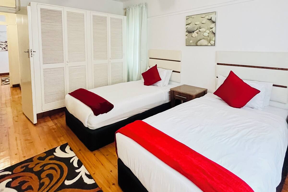 Habitación de hotel con dos camas, ventilador de techo, puerta blanca y alfombra estampada.