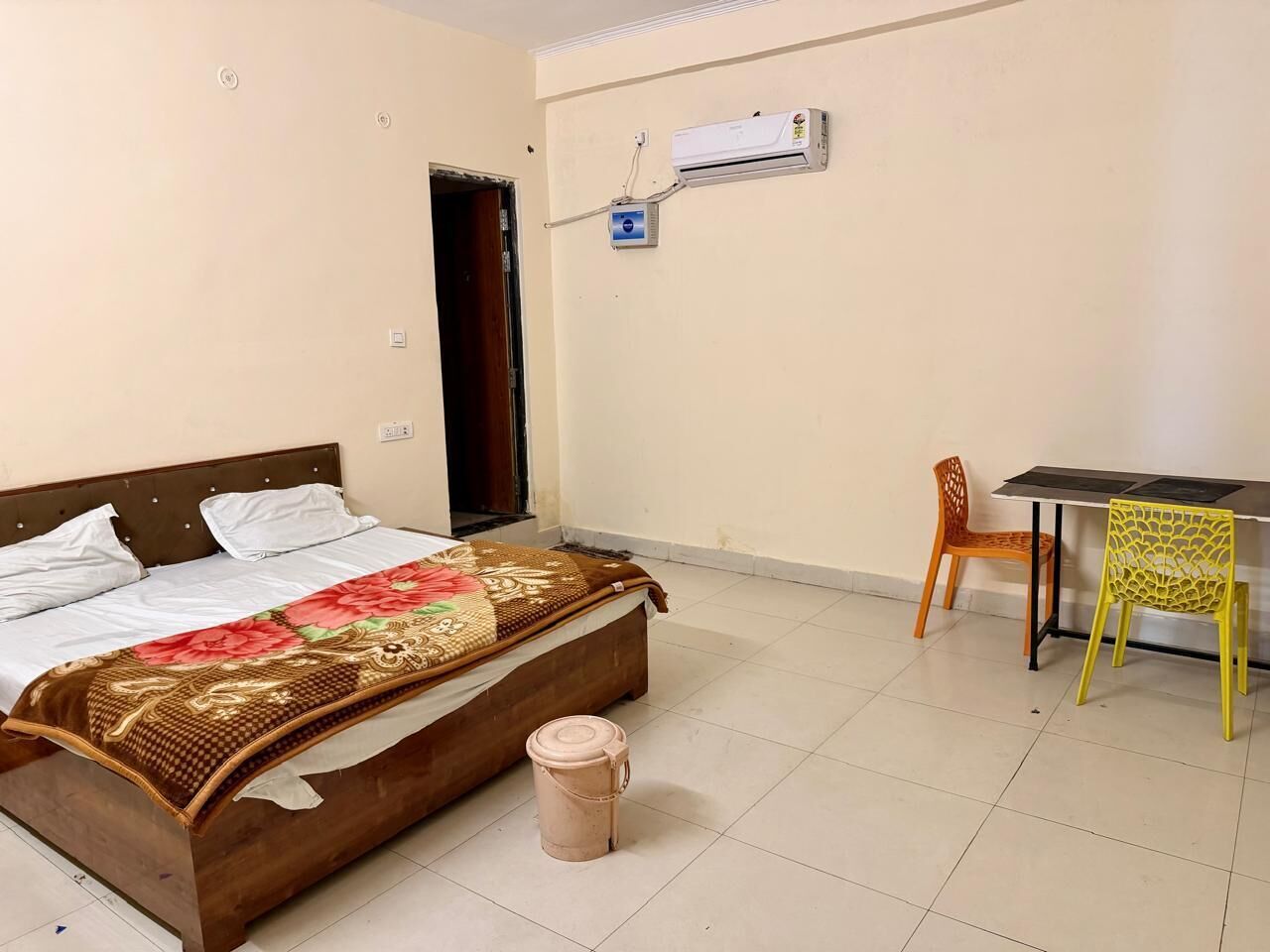 Een slaapkamer met een bed, een tafel, twee stoelen, een airconditioner en een afvalbak.