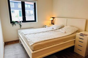 Una cama individual con cabecera blanca, una lámpara de noche, una mesita de noche pequeña y una ventana con vistas a edificios.