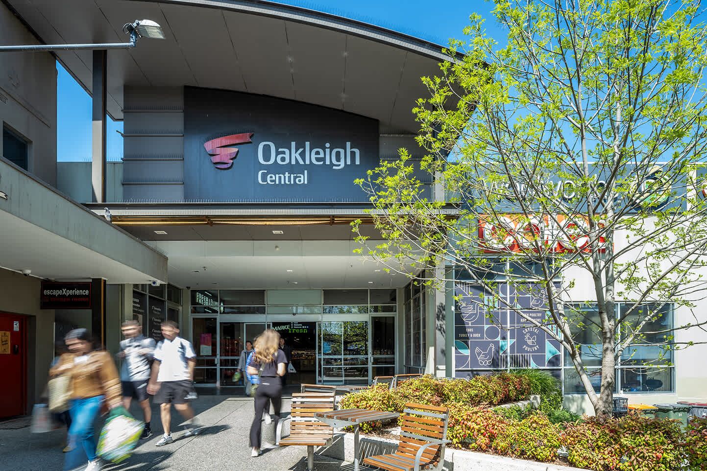 The exterior of Oakleigh Central with a modern design, including a large sign, benches, and a tree.