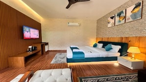 A modern hotel room with a wooden floor, a bed with white and blue bedding, a TV on the wall, a desk with a lamp, and artwork on the wall.