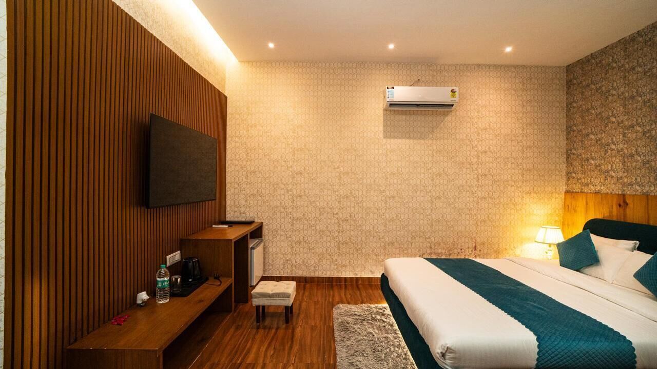 A hotel room with a wooden panel wall, a flat-screen TV, a bed with teal bedding, a bedside table with a lamp, and an air conditioning unit.
