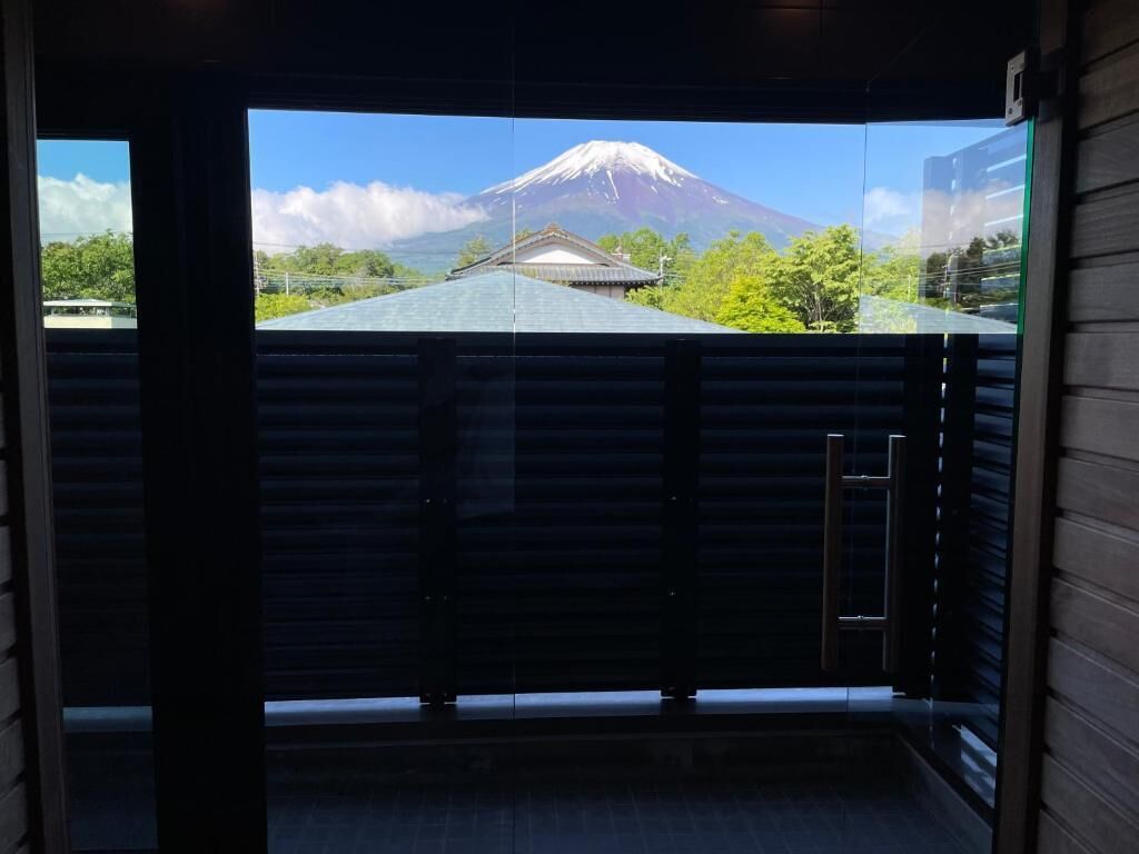 A view through a glass door of a building with a traditional Japanese roof, a mountain in the background, and a wooden fence.