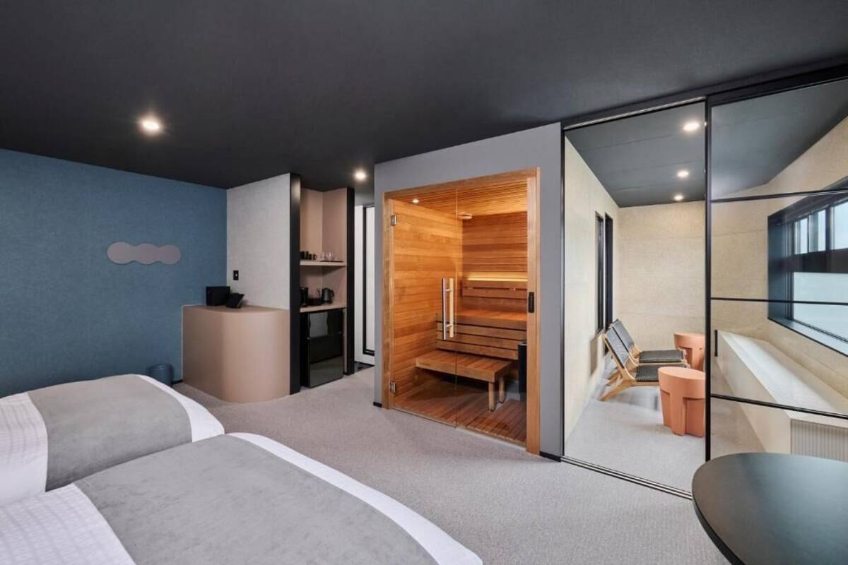 Una habitación de hotel moderna con dos camas, una sauna de madera, una zona de estar con una mesa y un ventanal grande.