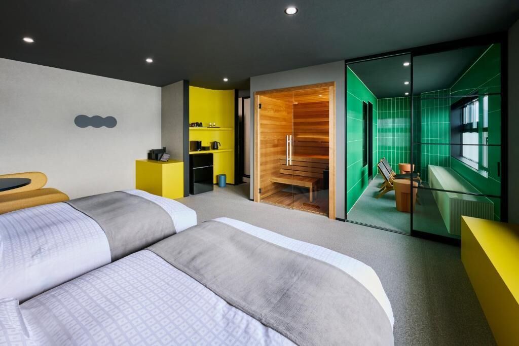 A modern hotel room with a large bed, a small desk, a seating area with a chair and table, and a view of a green-tiled room.