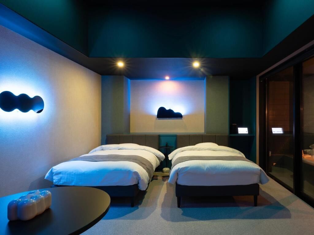 A hotel room with two beds, a small table, and a wall-mounted light fixture.