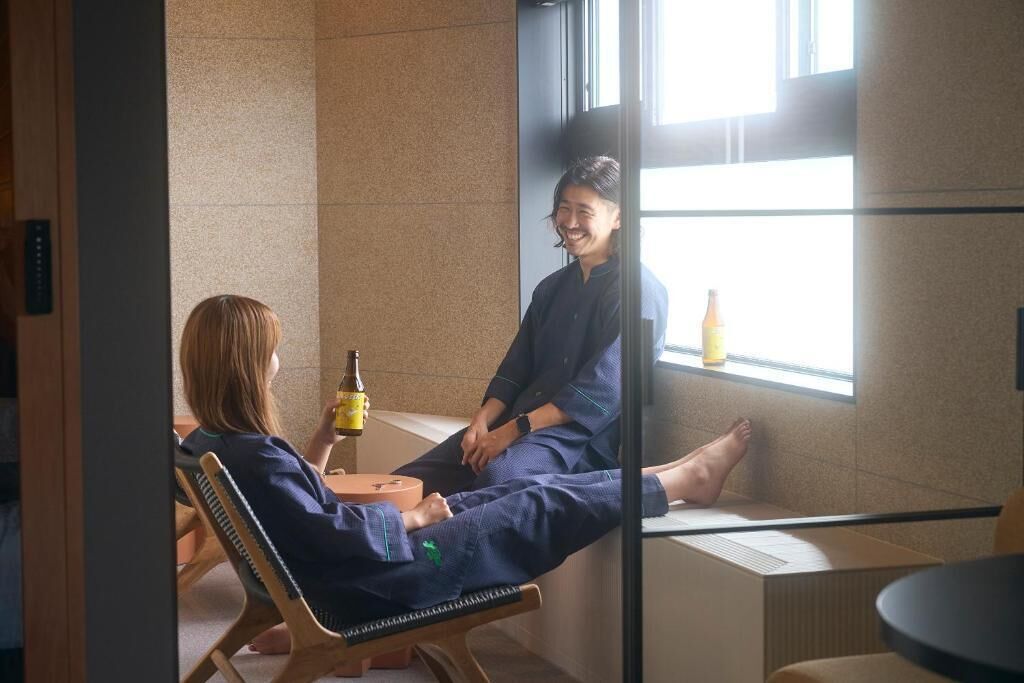 Una habitación con un ventanal grande, una botella en el alféizar y una persona sentada en una silla.