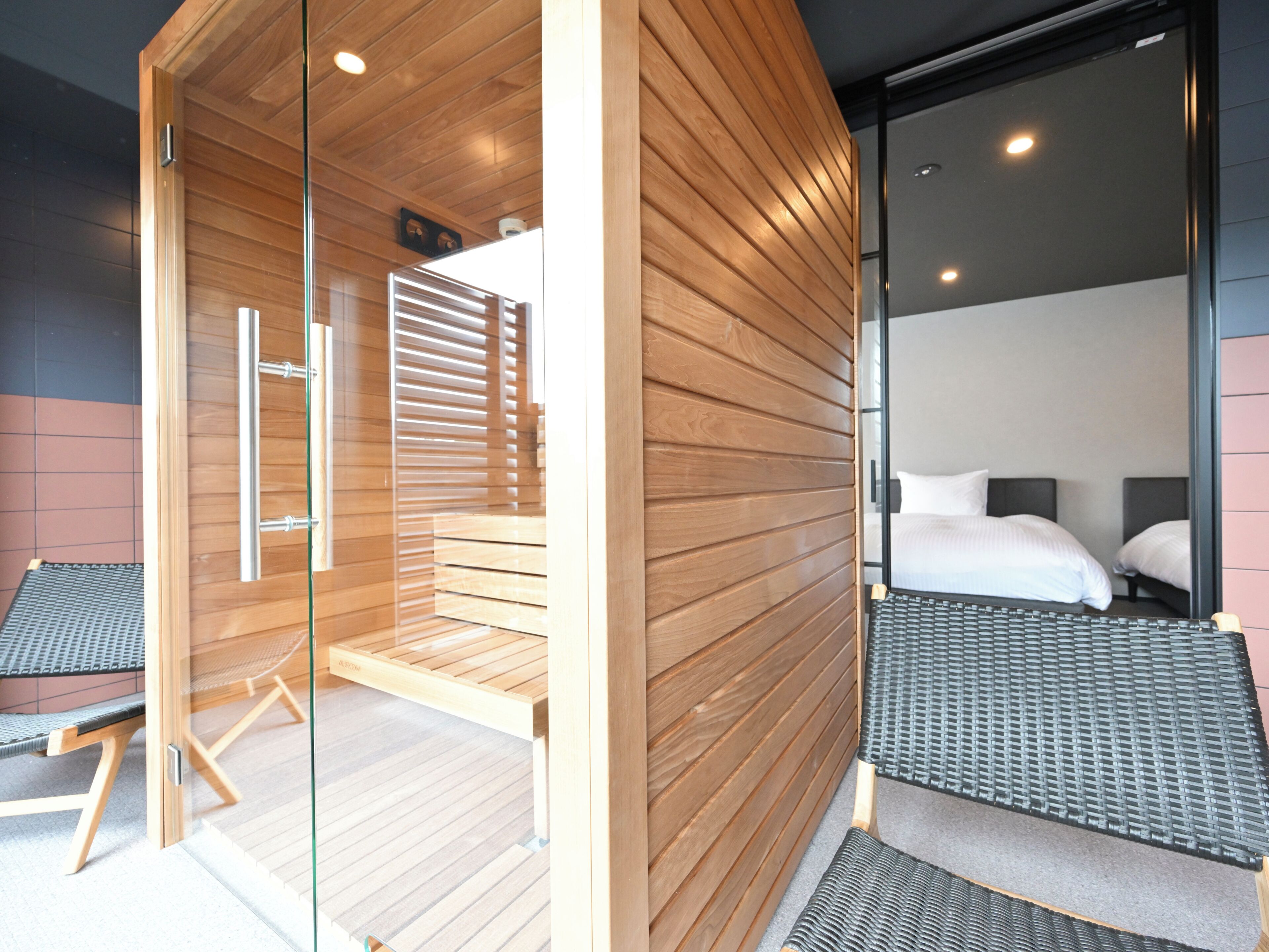 Une chambre d’hôtel moderne avec un intérieur en bois, une cloison en verre, un lit avec une literie blanche et un petit coin salon.
