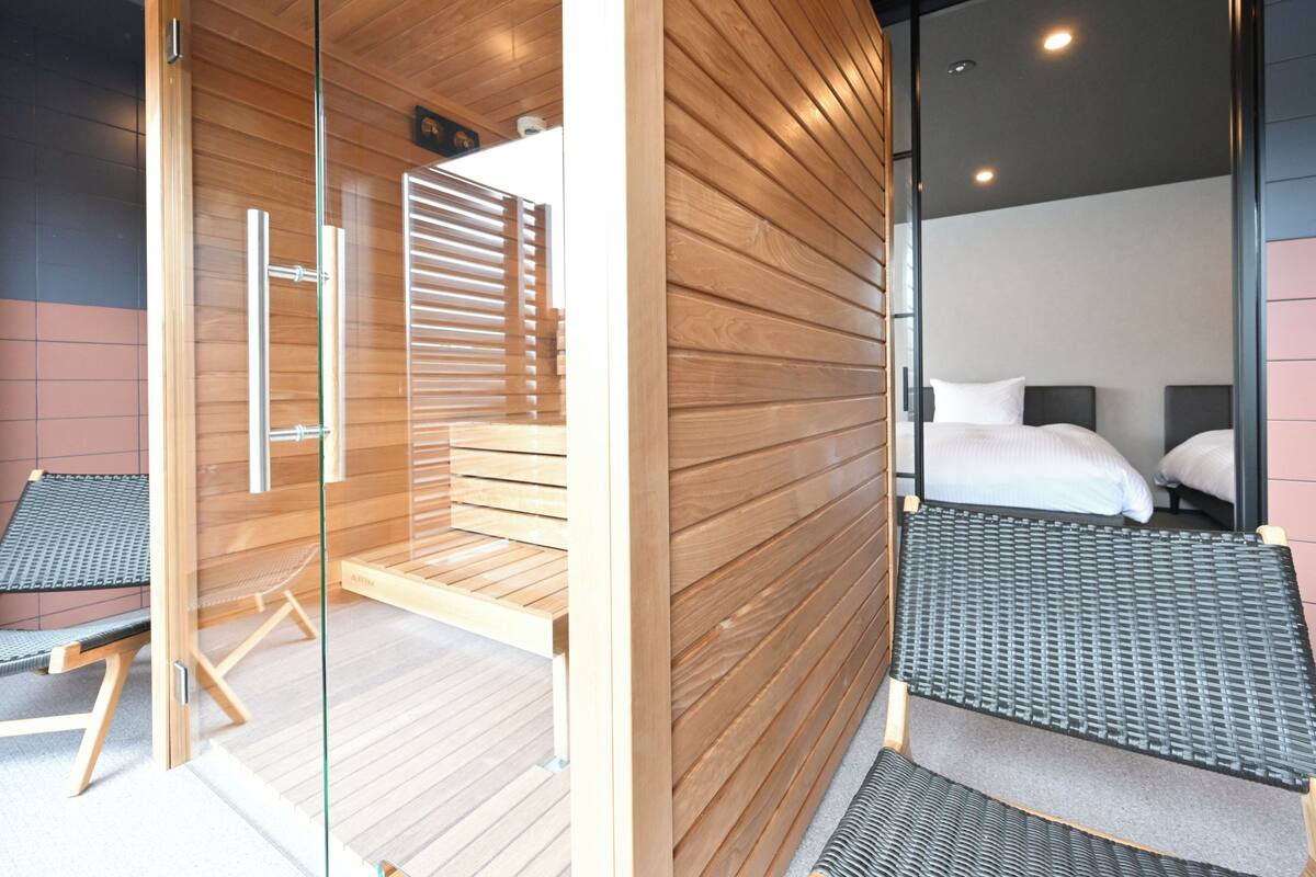 Una habitación de hotel moderna con interior de madera, un separador de vidrio, una cama con ropa de cama blanca y una pequeña zona de estar.