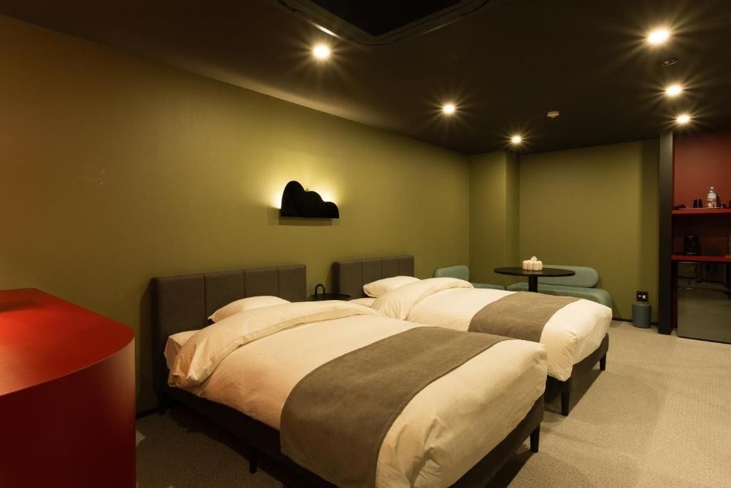 Una habitación de hotel con dos camas, una mesita pequeña, una silla roja y una repisa en la pared.
