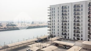 A high-rise building with multiple balconies, a parking area with cars, and a view of a body of water. - Bloomfields Unique 1BR Water's Edge2  (Abu Dhabi)