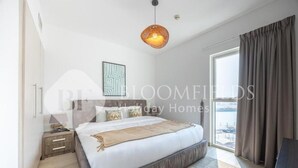 A bedroom with a large bed, a bedside table, a hanging light fixture, and a window with curtains. - Bloomfields Unique 1BR Water's Edge2  (Abu Dhabi)