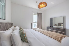 A modern bedroom with a bed, a dresser, a mirror, and a hanging light fixture. - Bloomfields 1BR in Water's Edge 2-Yas Island (Abu Dhabi)