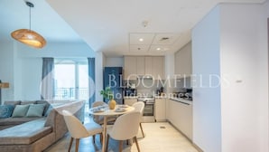 A modern living room with a sofa, dining table, and chairs. There is a kitchen area with cabinets and a window. - Bloomfields Unique 1BR Water's Edge2  (Abu Dhabi)