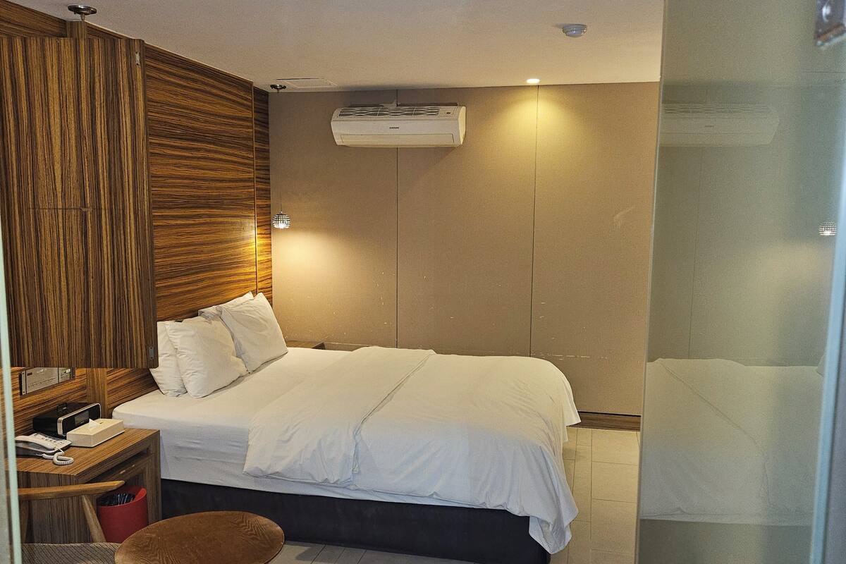 A modern hotel room with a bed, bedside table, lamp, and an air conditioning unit.