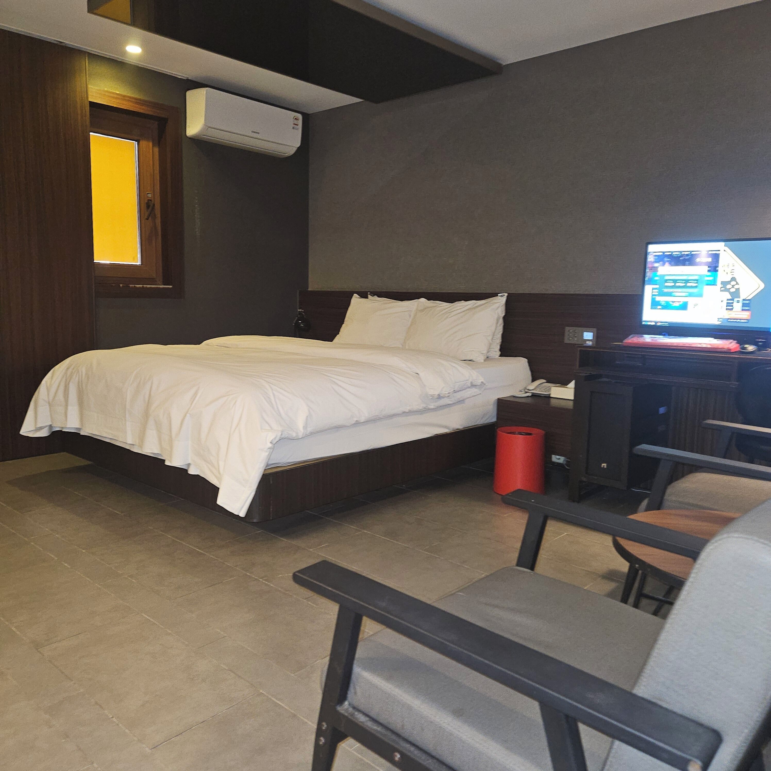 Une chambre d’hôtel comprenant un lit, un bureau avec un ordinateur, une chaise, une poubelle rouge et un appareil de climatisation.