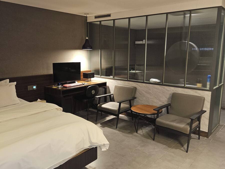 Habitación de hotel con cama, escritorio con televisor, dos sillas, una mesita y un ventanal grande con persianas.