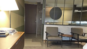 Une chambre d’hôtel avec un bureau en bois, une lampe, une chaise, une petite table et une grande fenêtre avec des stores.