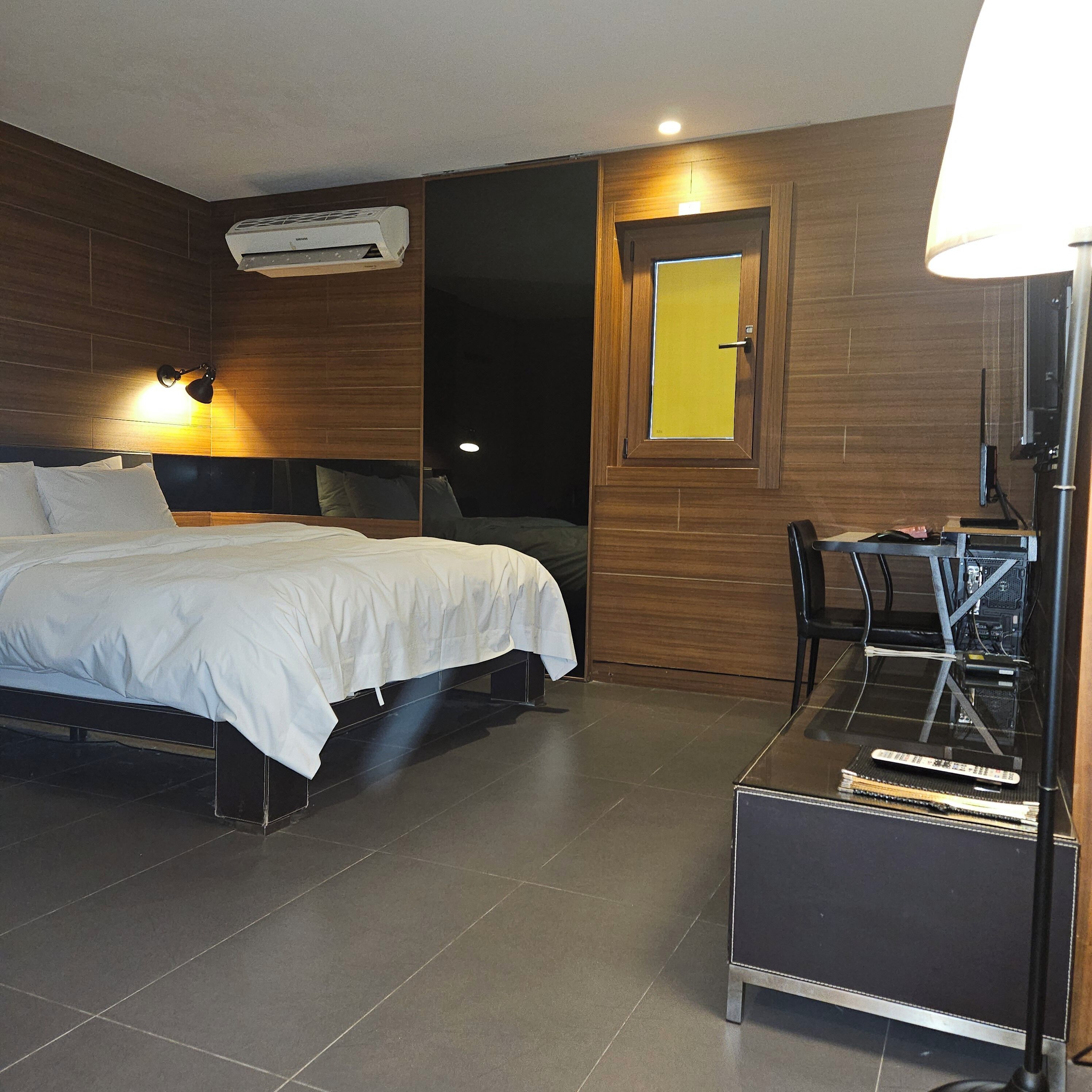 Una habitación de hotel con cama, escritorio y silla, unidad de aire acondicionado y una puerta con marco amarillo.