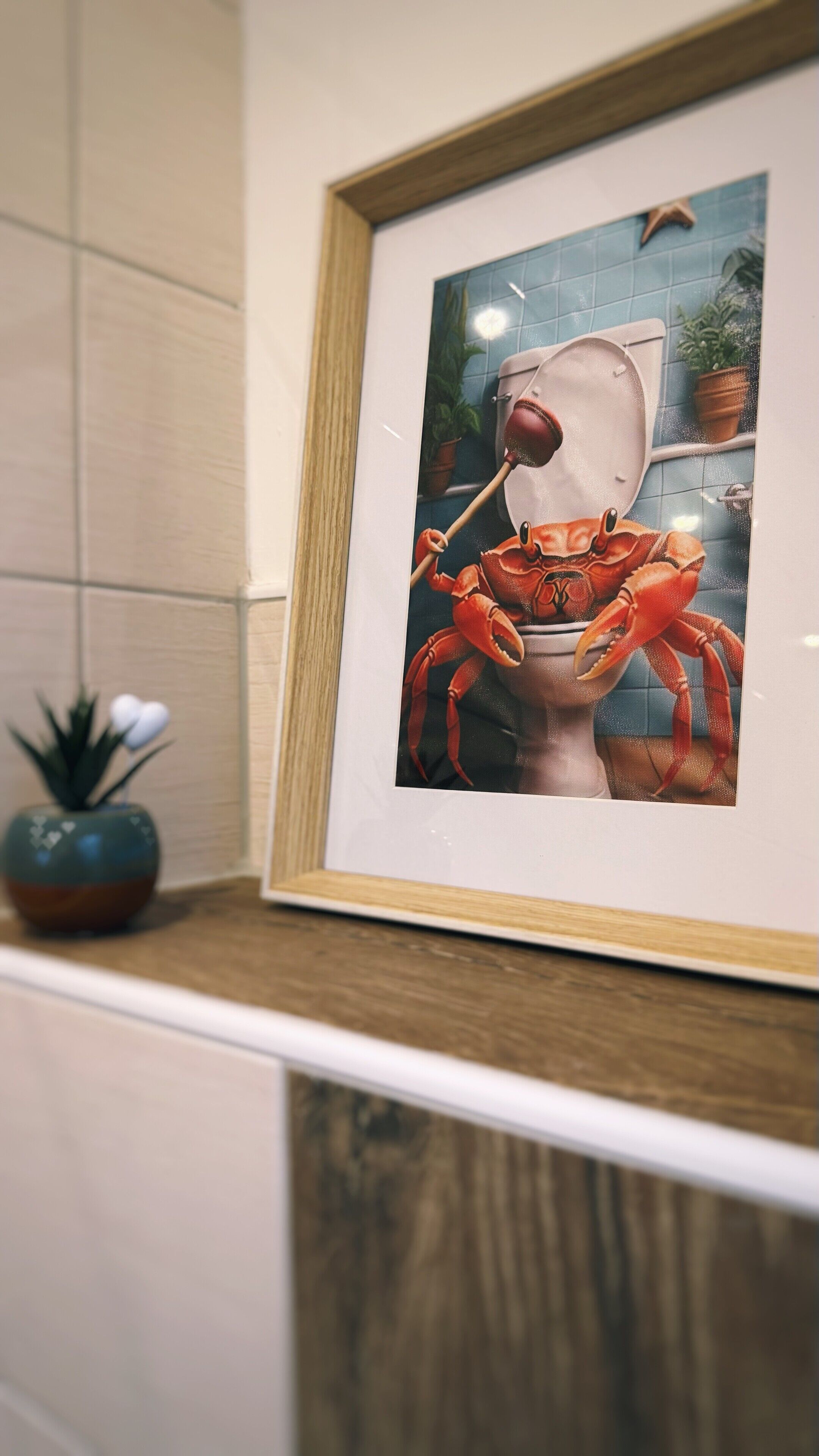 Een lijst met een afbeelding van een krab op een toiletbril, een potplant en een houten plank.