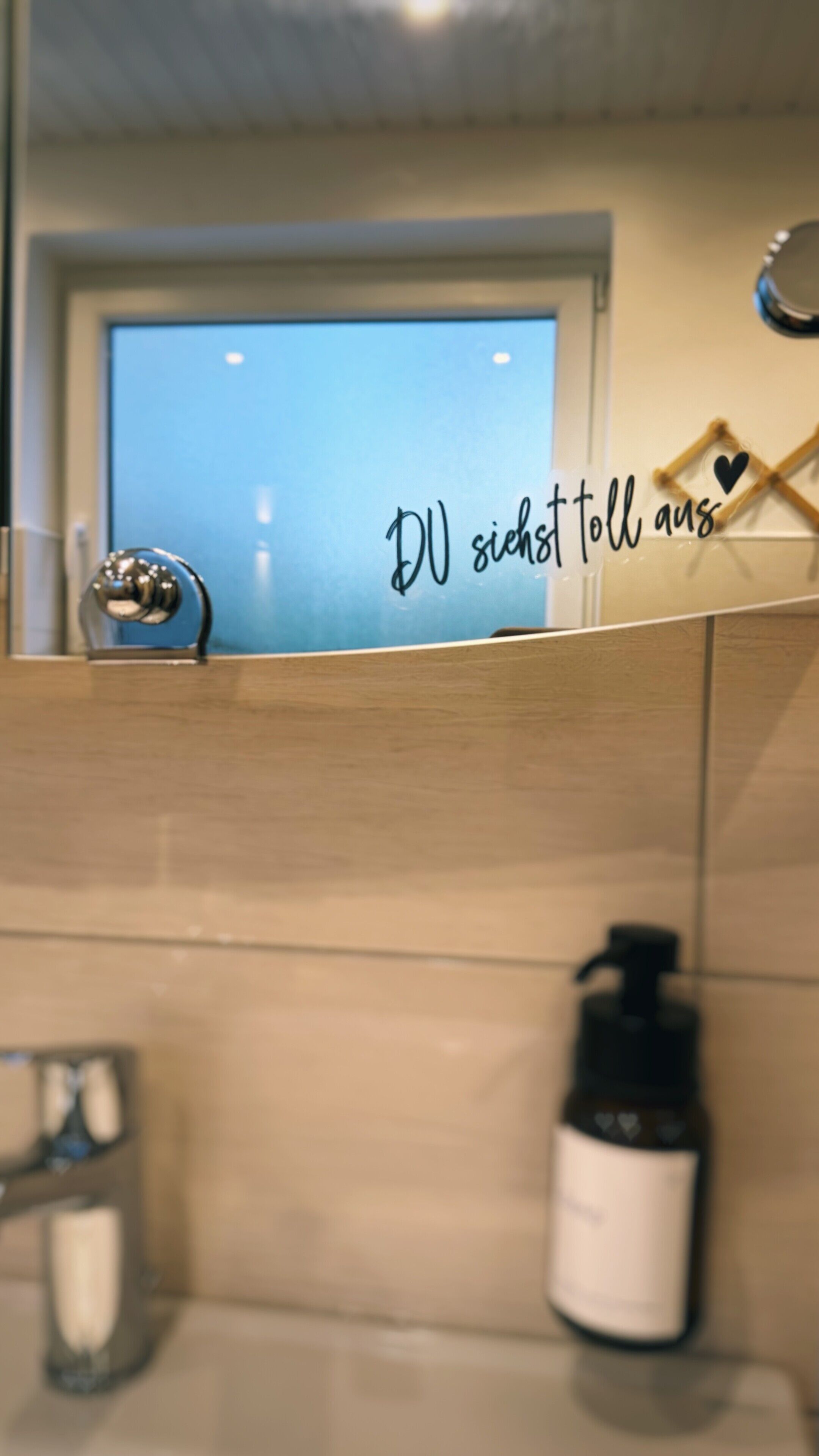Et badeværelse med et spejl, håndspritdispenser og en væg med en tysk inskription.