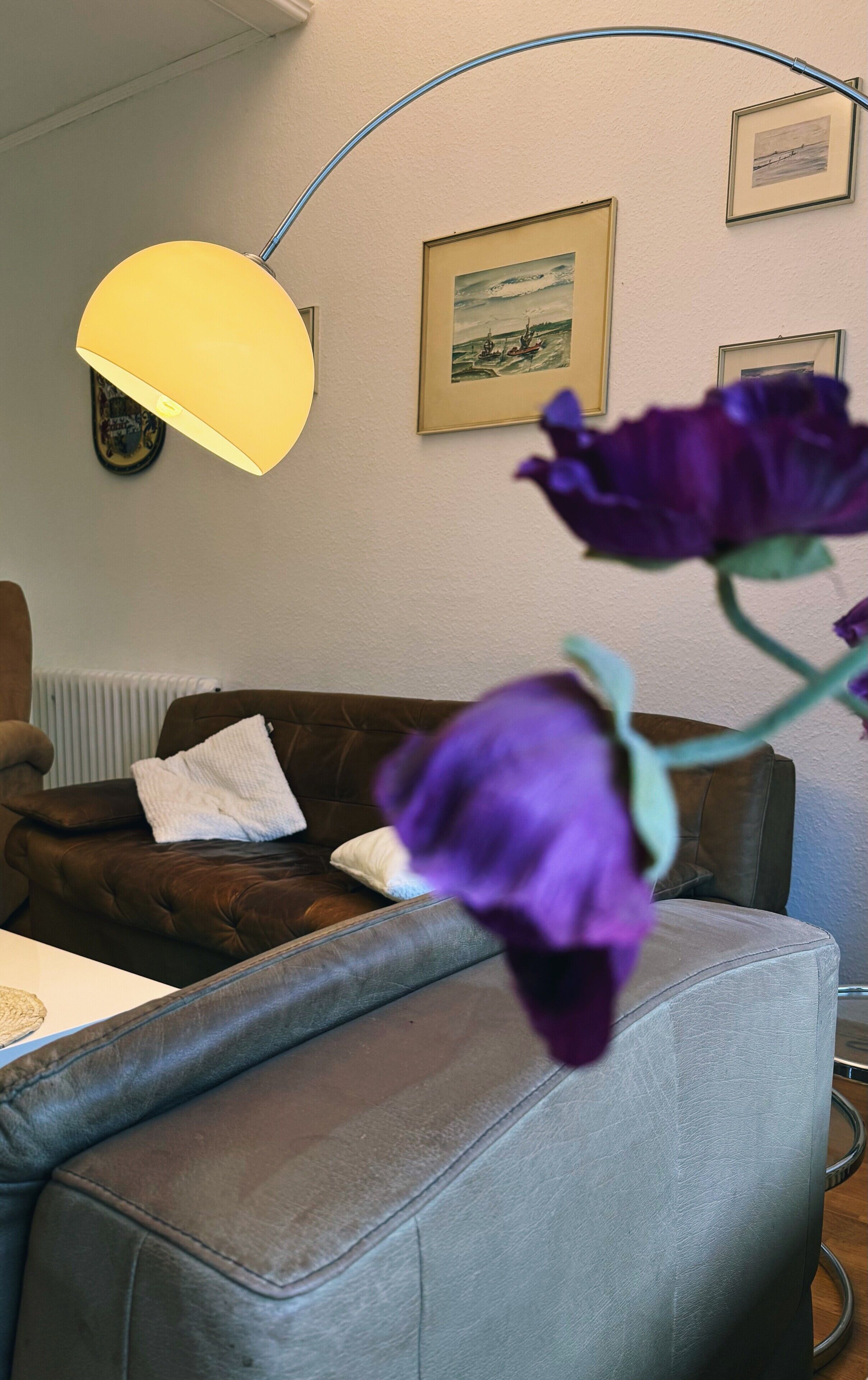 Una sala de estar con un sofá de cuero marrón, una lámpara de pie amarilla y flores moradas en primer plano.