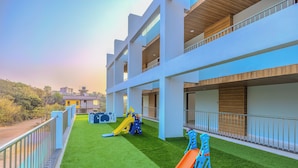 Un edificio moderno con varios balcones, una zona de juegos infantiles con toboganes y césped artificial.
